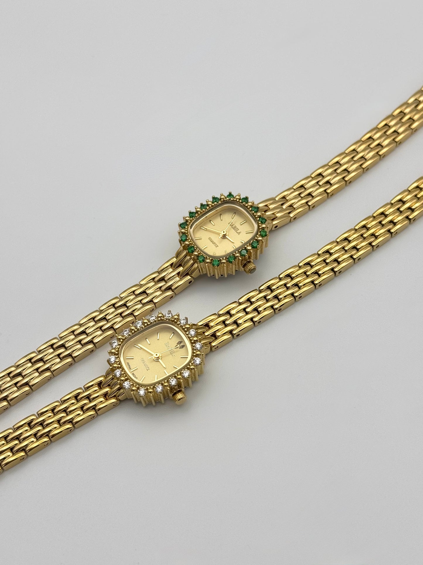 A gold plated vintage styled watch with a floral pattern on the band and a gold dial. Green and white gemstones