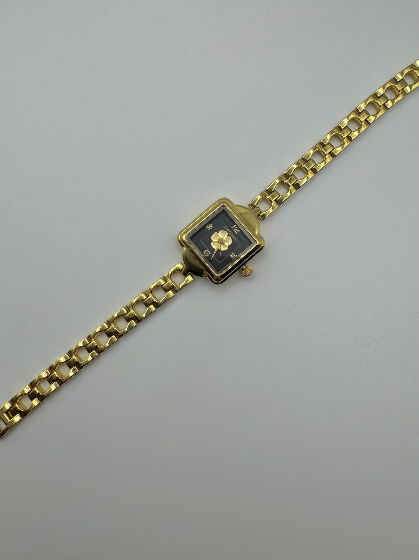 Gold wristwatch with a square face and centered flower on a black dial. Minimal vintage styled