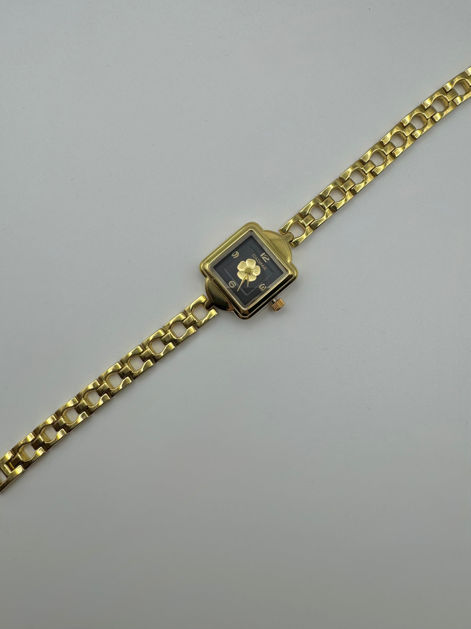 Gold wristwatch with a square face and centered flower on a black dial. Minimal vintage styled