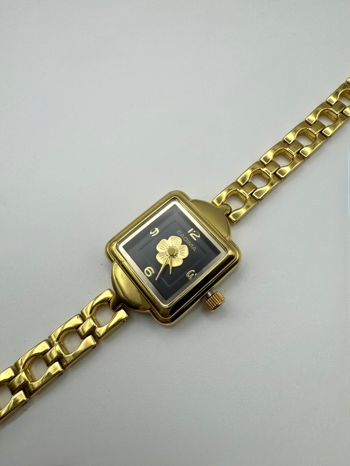 Gold wristwatch with a square face and centered flower on a black dial. Minimal vintage styled
