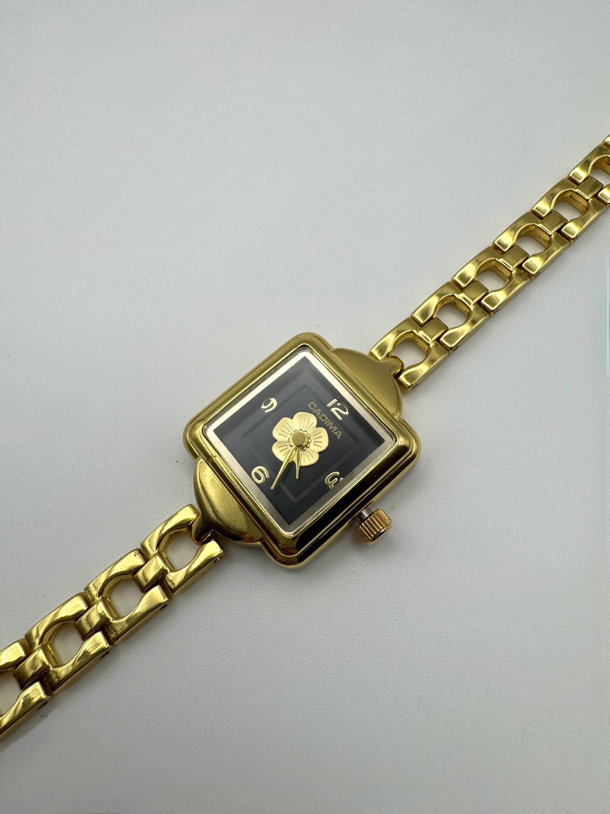 Gold wristwatch with a square face and centered flower on a black dial. Minimal vintage styled