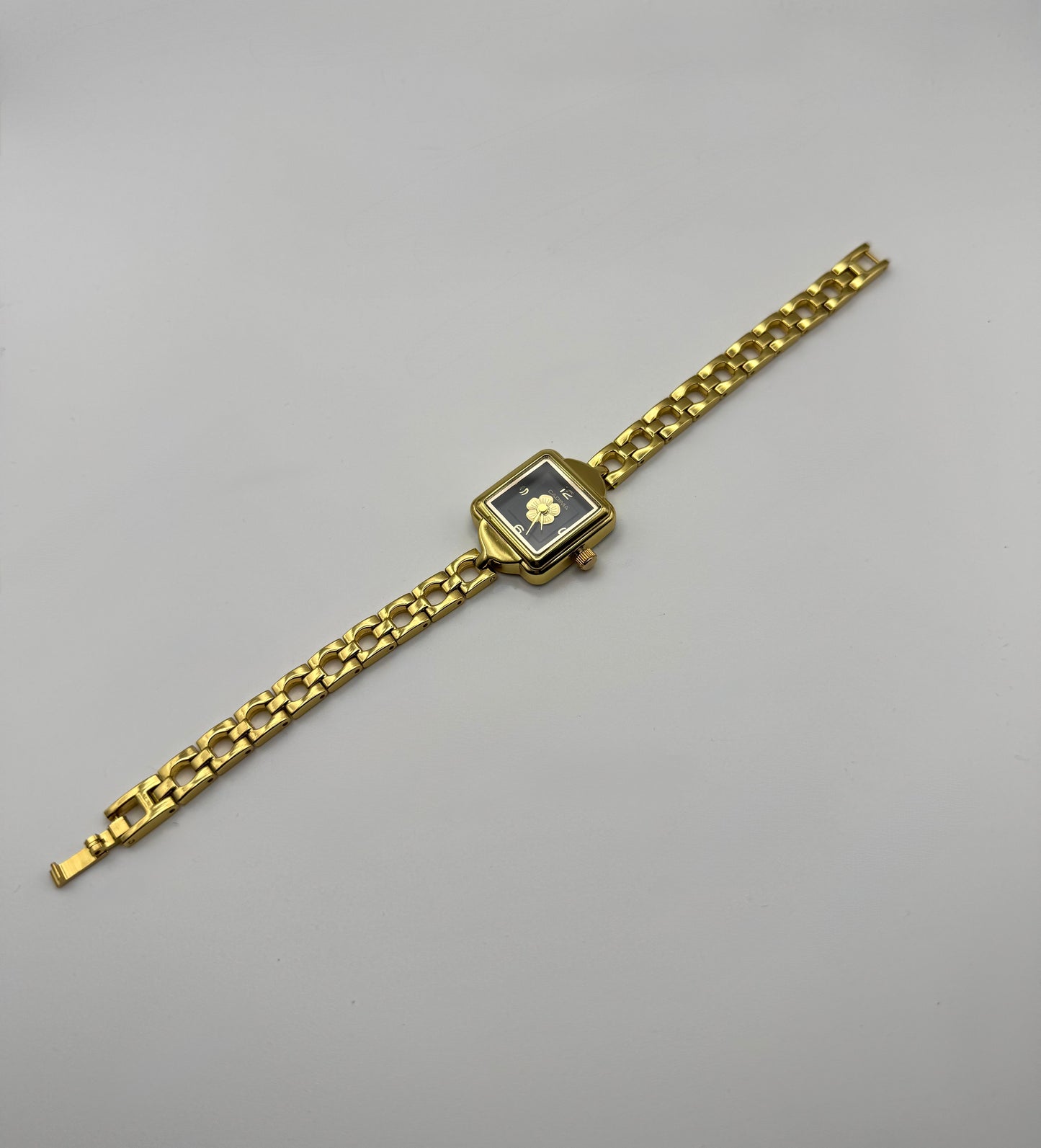 Gold wristwatch with a square face and centered flower on a black dial. Minimal vintage styled